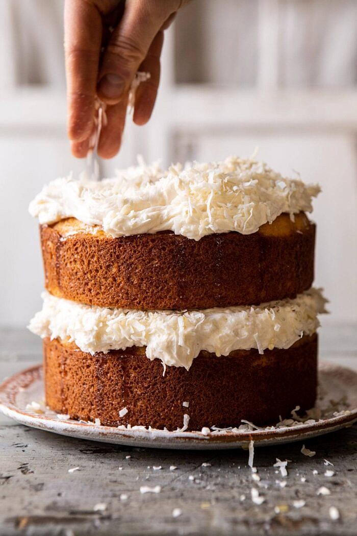 Lemon Coconut Naked Cake with Whipped Vanilla Buttercream being sprinkle with coconut and hand in photo 